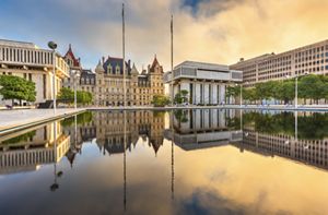 This historic building that serves as New York's state capitol is reflected in a plaza pool at sunrise. It is surrounded by other historic and modern government buildings.