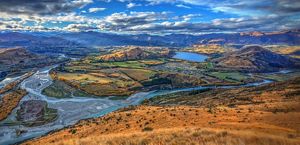 Aerial view of Lake Hayes, New Zealand, showing a meandering river winding around hills with mountain ranges in the distance.