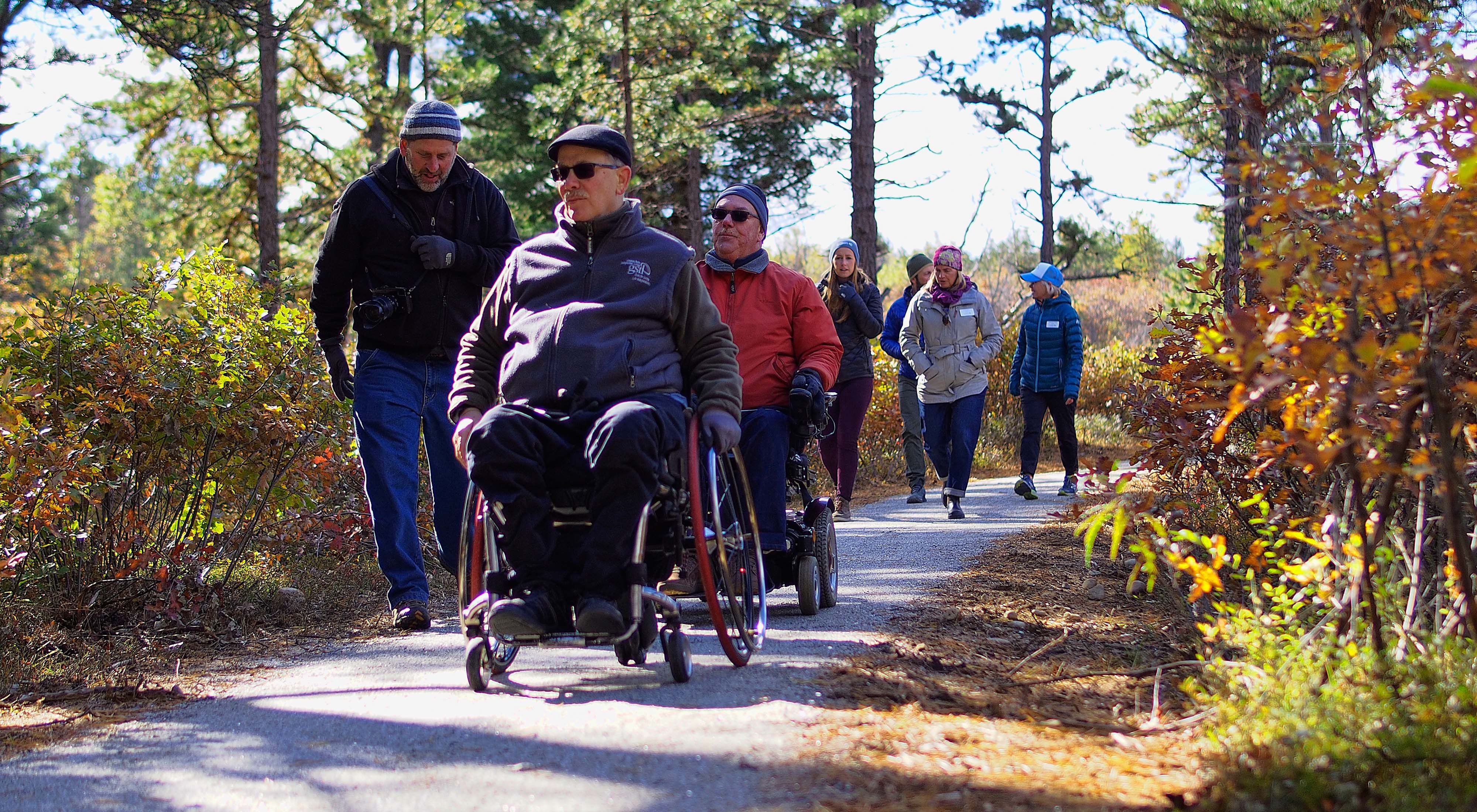 A man in a wheelchair with a group of followers on an accessible trail in the Ossipee Pine Barrens