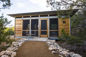 A gravel trail leads to a wooden cabin with a metal roof surrounded by dense brush.