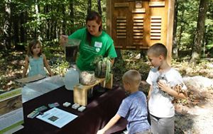 A woman stands at a table at an outdoor visitors center. She holds a pitcher full of dirty water showing three children a water quality testing experiment.