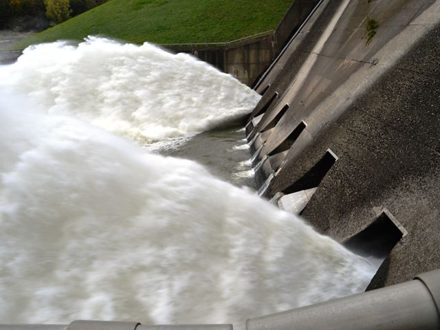 White water rushes through two square openings at the bottom of a concrete dam. The water fans out in high jets from the speed of its flow.