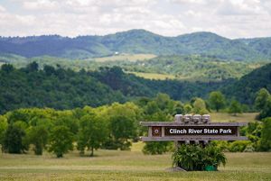 A large sign reading Clinch River State Park welcomes visitors. Rolling tree covered hills rise behind the sign toward a tall mountain ridge that lines the horizon.