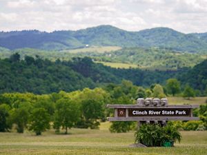 A large sign reading Clinch River State Park welcomes visitors. Rolling tree covered hills rise behind the sign toward a tall mountain ridge that lines the horizon.