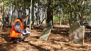 Man kneels with pen and paper in front of old tombstone in wooded area.