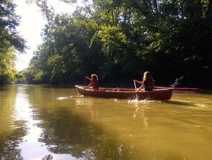 Two women paddling in a canoe on a muddy, tree-lined river.