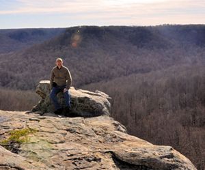 A man stands against a rock in front of a vast valley.