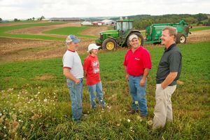 Four people standing in a farm field talking; a large green tractor is in the background. 