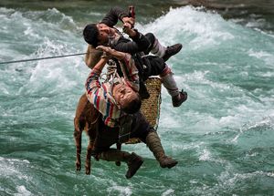 Two people use a rope and pulley system to cross a raging river with a basket and an animal.