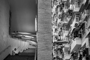 A black and white view of buildings in Hong Kong. On one side is a person sitting in a stairwell alone, and on the other side is dozens of balconies in a tall building.