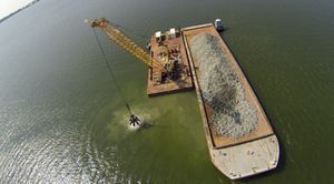 Aerial view looking down on a floating baroe in the Piankatank River. A large crane is being used to scoop up chunks of granite rock from a pile on the barge and place them on a new oyster reef.