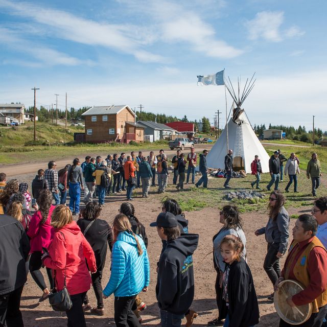 Dozens of people form a large circle, with a teepee in the background.