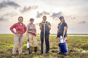 Four women stand in an expanse of green wetlands holding tools.