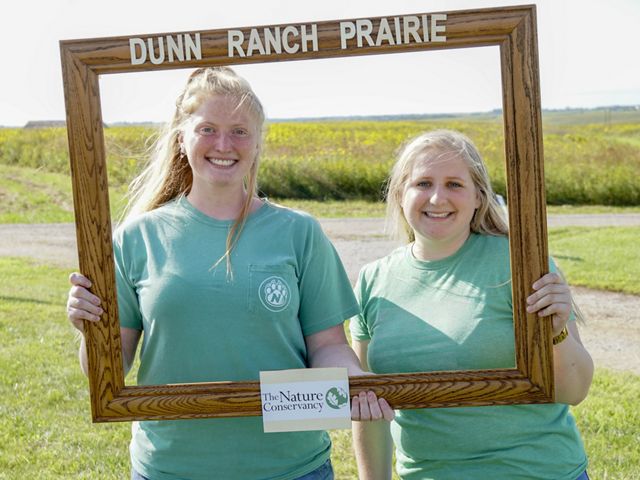 Two women smile and pose while holding up a wooden frame labeled 'Dunn Ranch Prairie.'