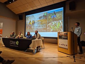 Several people sit at a panel conference table while another person stands at a podium; a PowerPoint presentation is projected on a large screen behind them.