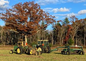 A tractor and hay baler, both green and yellow, rest in an open hayfield in front of a large oak tree with autumn foliage.