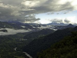 Aerial view of heavily forested mountains in Ecuador, with a river winding through a valley among the mountains and dramatic clouds overhead.