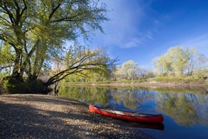 A red canoe rests on the shore of a calm river lined with trees.