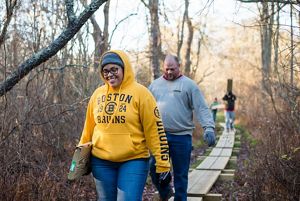 Two people carry lumber along a boardwalk, walking toward the camera in a wooded area.