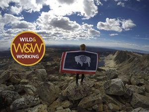 Girl with Wyoming state flag stands on mountain under cloudy blue skies.