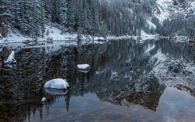 Snow-covered trees surround a lake that reflects Hallet Peak during an early season snowfall in Rocky Mountain National Park.