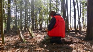 A man kneels on the ground in front of two weathered gravestones, recording the information on the markers.