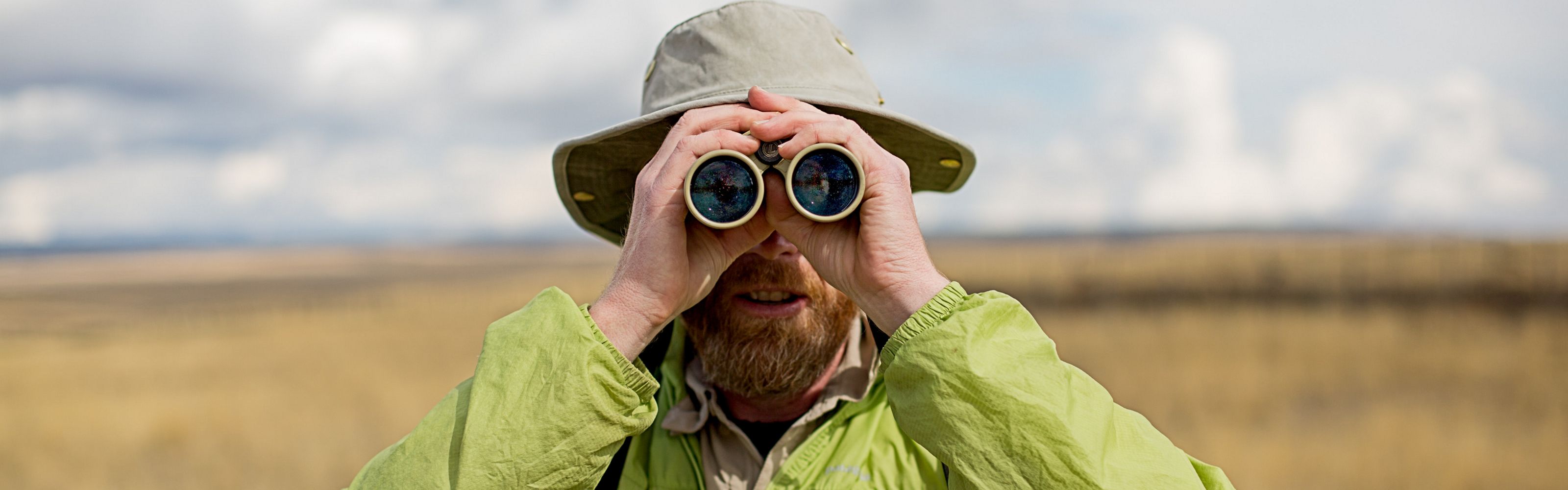 Person wearing hat and looking through binoculars while facing the camera.