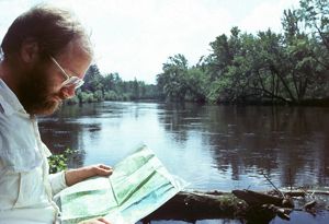 In the foreground, a bearded man looks at a map in front of a lake. 