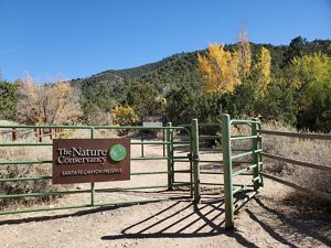 View of the entrance of a nature preserve, with a sign reading The Nature Conservancy Santa Fe Canyon Preserve and pine trees in the distance.