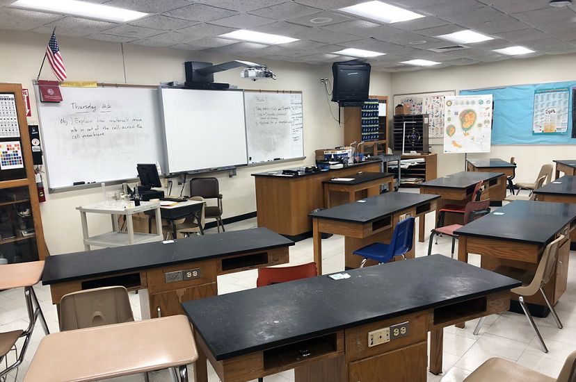 A high school classroom is filled with educational posters, a long white board and desks with electrical outlets.