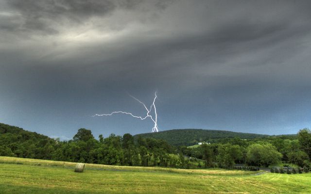 Lightning forks from a heavy gray sky arcing behind a green mountain ridge. A line of trees runs between two open fields.