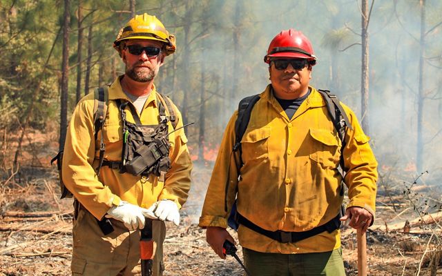Gesse Bullock of the Alabama-Coushatta Tribe of Texas and Shawn Benedict of TNC Texas stand in their yellow protective gear, holding fire tools as a prescribed burn blazes in the forest behind them.