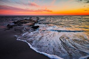 A wave foams on the shore as the sun sets over the ocean.