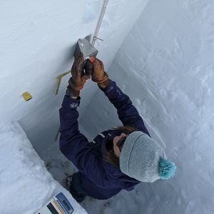 A woman in cold-weather gear measures snow as she stands in a large square hole. 