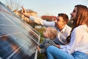 Man shows his family the solar panels on the plot near the house during a warm day. Young woman with a kid and a man in the sun rays look at the solar panels. 