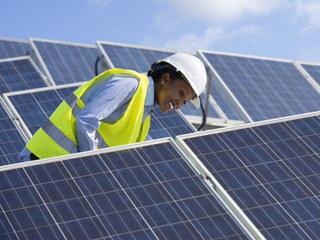 A worker in a hard hat stands between rows of solar panels.