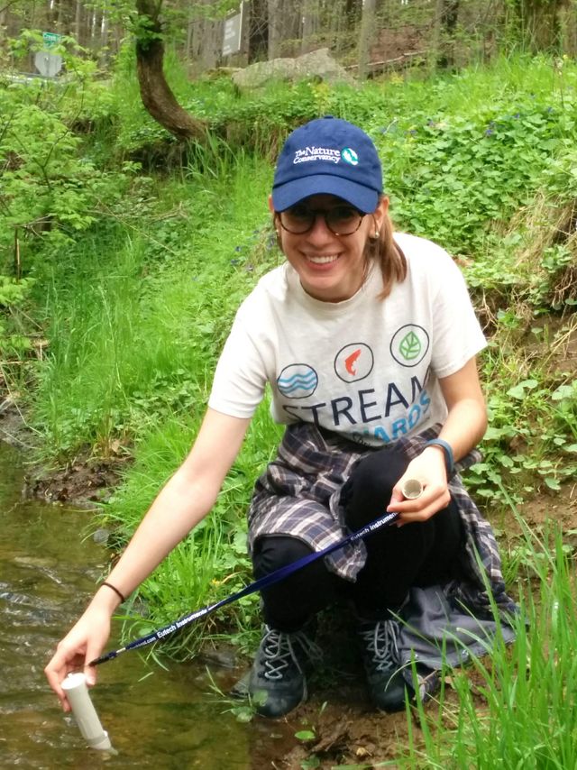 A smiling woman crouches down at the edge of a stream. She is holding one end of a white tube used to sample water quality.