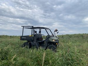 A man stands next to a UTV and sprays invasive plants in a wide field.