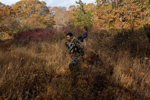 man in camo walks through grassy field holding compound bow.