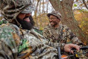 A close-up image of 2 men of color wearing camo and laughing together, sitting in the woods.