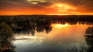 The sun breaks over the horizon and orange skies are reflected in a pond below, ringed by pine trees. 