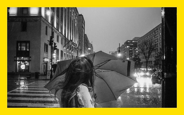 Black and white photo of a person holding an umbrella against a dark and rainy city street.