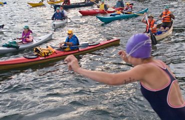 A person on the right hand side of the frame is mid-jumping into the water. They are wearing a purple swim cap, goggles, and a one-piece swimsuit. Many kayakers are onlooking.