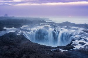 Tidal waters rushing into a large hole in black coastal rocks surrounded by blue sea and purple skies.