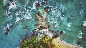 Waves break over rocks on the shoreline in an aerial image over a small spit of rocky and sandy land.