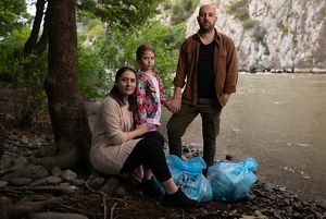 A family stand on the banks of a river with bags of items for a picnic.