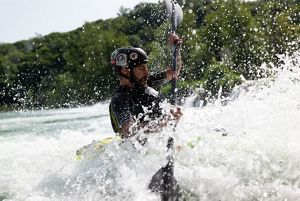 A kayaker wearing a helmet navigates through splashes and whitewater rapids on a sunny day.