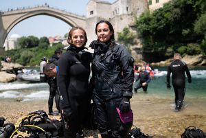 Two divers in wet suits smile and pose on a river bank with an old arched stone bridge in the background.
