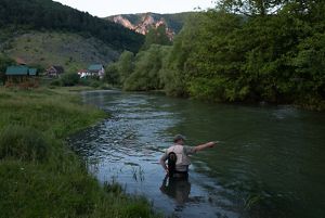 A fisherman stands in a slow bend of a river and casts a rod around dusk with farms in the background.