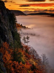 Sunrise over a fog-covered forest in autumn, showing vibrant orange and yellow foliage with rugged cliffs on the left.